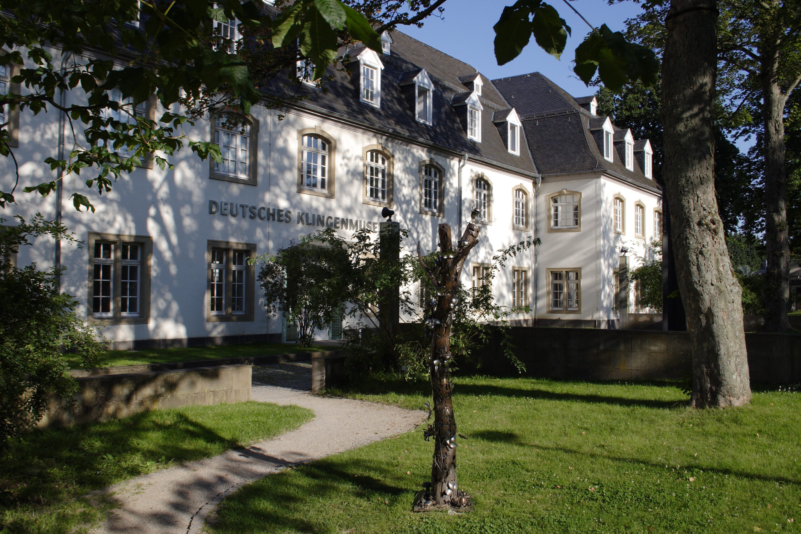 Ein weißes Museumsgebäude mit der Aufschrift „Deutsches Klingenmuseum“, umgeben von Bäumen und einer gepflegten Grünanlage. Im Vordergrund führt ein geschwungener Weg zu einer Skulptur im Garten.