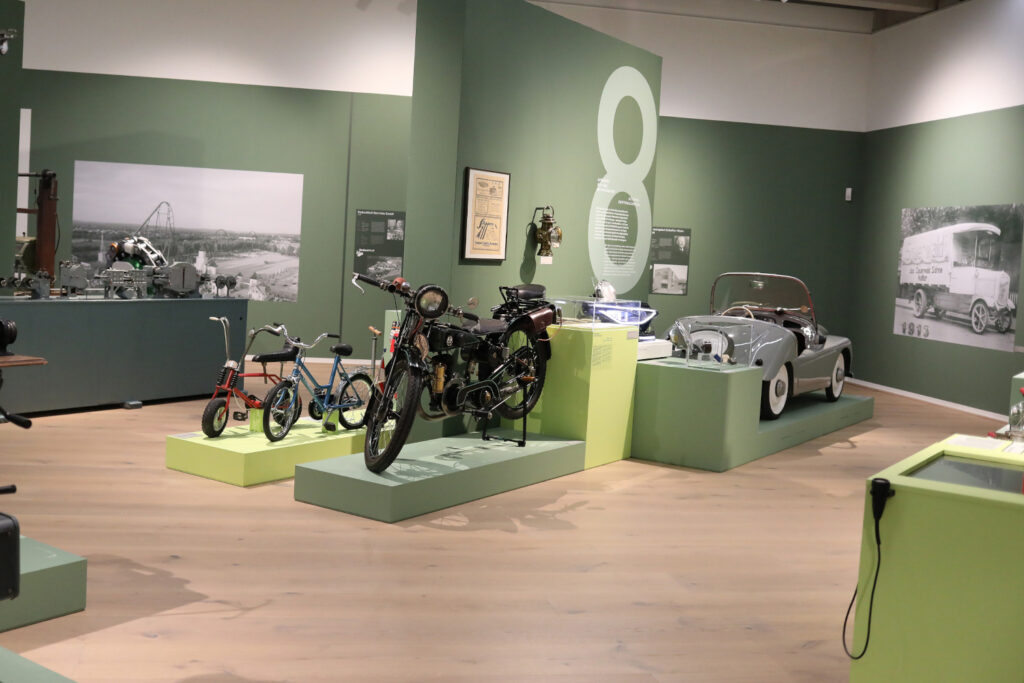 Ausstellungsraum mit historischen Fahrzeugen und Exponaten. Im Vordergrund stehen ein schwarzes Motorrad sowie zwei Kinderfahrräder auf Podesten. Rechts ist ein silberner Oldtimer-Cabrio ausgestellt. An den grün gestalteten Wänden befinden sich großformatige historische Fotografien, Informationstafeln und eine überdimensionale Zahl „8“, die auf einen Themenbereich der Ausstellung hinweist.