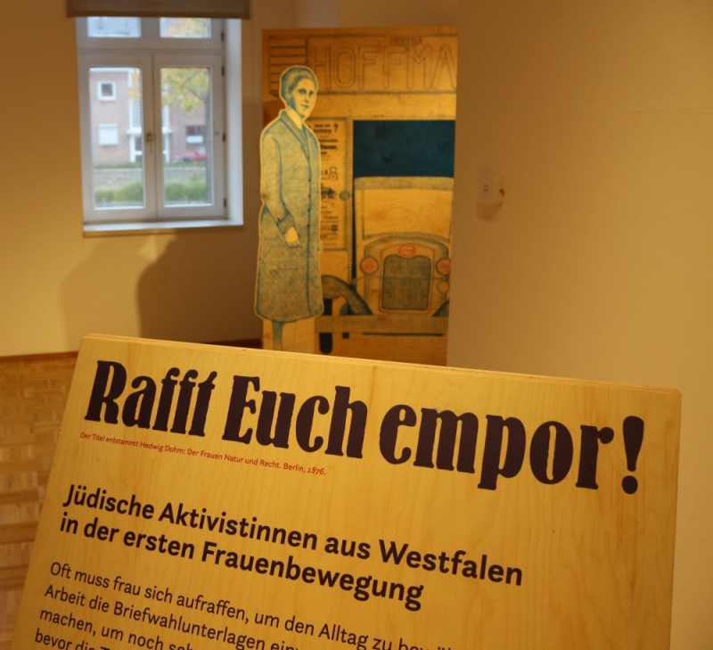 Blick in eine Museumsausstellung mit einer schräg aufgestellten Informationstafel im Vordergrund. Darauf steht der Titel „Rafft Euch empor!“ sowie der Untertitel „Jüdische Aktivistinnen aus Westfalen in der ersten Frauenbewegung“. Im Hintergrund sind eine lebensgroße Figur einer Frau als Aufsteller, ein historisches Fahrzeugmotiv und ein Fenster zu sehen. Die warm beleuchtete Ausstellung vermittelt einen historischen Kontext zur Frauenbewegung und jüdischen Aktivistinnen.