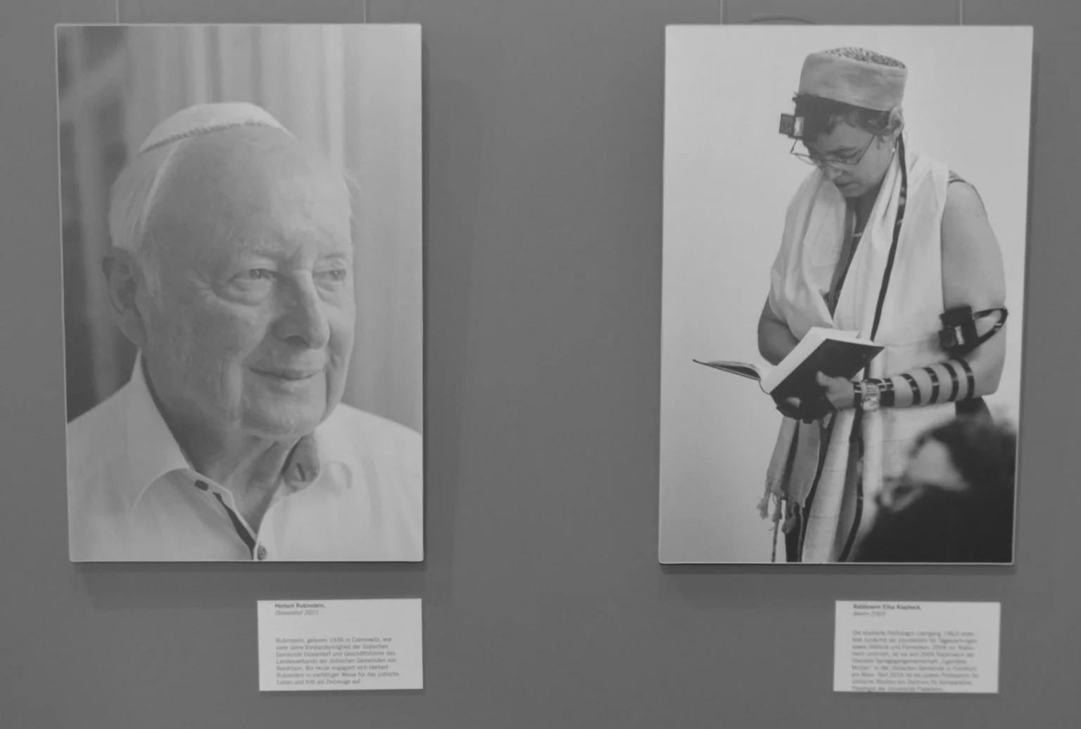 Auf dem Bild sind zwei Fotos abgebildet. Im musealen Kontext sind die Fotos auf einer dunkelgrauen Wand angebracht, darunter sind Texttafeln. Beide Fotos sind schwarz-weiss, auf dem linken Bild Sieht man einen älteren Mann, er wird als Herbert Rubinstein benannt. Auf dem rechten Bild ist die Rabbinerin Elisa Klapheck im Gebet zu sehen.
