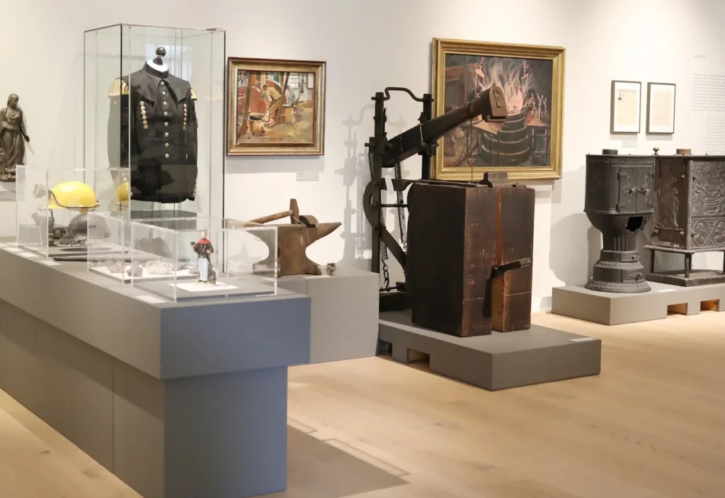 Ein heller Museumsraum mit einer Ausstellung historischer Objekte. Im Vordergrund stehen Vitrinen mit einem Bergmannshelm, einer Uniform, Werkzeugen und einer kleinen Figur. Daneben sind ein Amboss, eine große historische Maschine aus Holz und Metall sowie zwei verzierte gusseiserne Öfen zu sehen. An den Wänden hängen Gemälde mit Szenen aus der Metallverarbeitung sowie Informationstafeln zur Ausstellung.