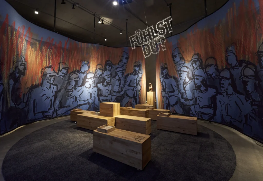 Runder Ausstellungsraum mit einer umlaufenden Illustration von Menschen in Uniform vor einem flammenartigen Hintergrund. In der Mitte laden hölzerne Sitz- und Medienstationen unter dem Schriftzug „FÜHLST DU?“ zum Verweilen und Informieren ein.