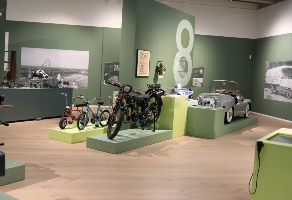 Ausstellungsraum mit historischen Fahrzeugen und Exponaten. Im Vordergrund stehen ein schwarzes Motorrad sowie zwei Kinderfahrräder auf Podesten. Rechts ist ein silberner Oldtimer-Cabrio ausgestellt. An den grün gestalteten Wänden befinden sich großformatige historische Fotografien, Informationstafeln und eine überdimensionale Zahl „8“, die auf einen Themenbereich der Ausstellung hinweist.