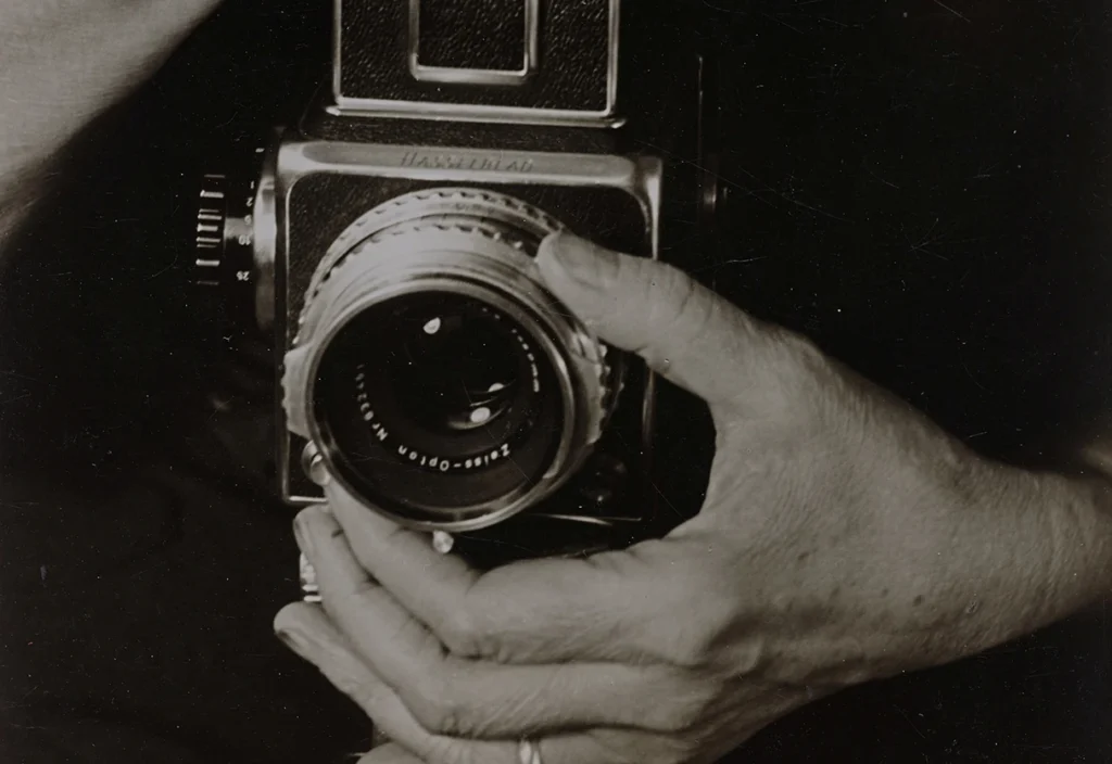 Schwarz-Weiß-Fotografie einer Mittelformatkamera, die von zwei Händen gehalten und bedient wird. Eine Hand umfasst den Kamerasucher von oben, die andere greift von unten an das Objektiv, als würde sie den Fokusring einstellen. Die Kamera ist zentral im Bild platziert, der Hintergrund ist dunkel und lenkt den Blick auf die Hände und das technische Gerät.