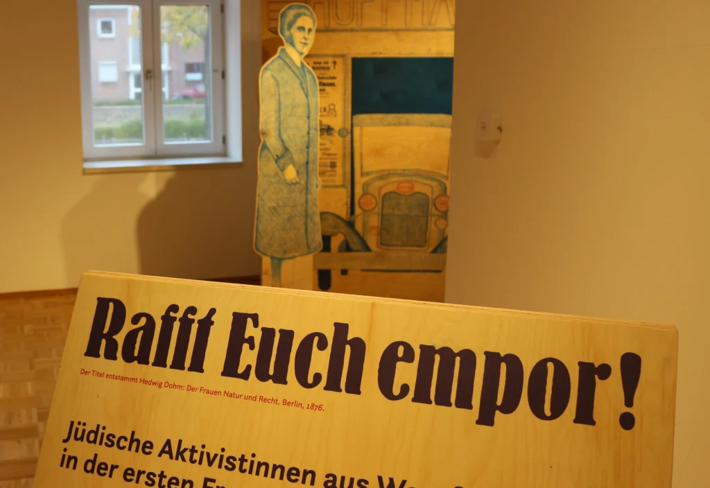 Blick in eine Museumsausstellung mit einer schräg aufgestellten Informationstafel im Vordergrund. Darauf steht der Titel „Rafft Euch empor!“ sowie der Untertitel „Jüdische Aktivistinnen aus Westfalen in der ersten Frauenbewegung“. Im Hintergrund sind eine lebensgroße Figur einer Frau als Aufsteller, ein historisches Fahrzeugmotiv und ein Fenster zu sehen. Die warm beleuchtete Ausstellung vermittelt einen historischen Kontext zur Frauenbewegung und jüdischen Aktivistinnen.