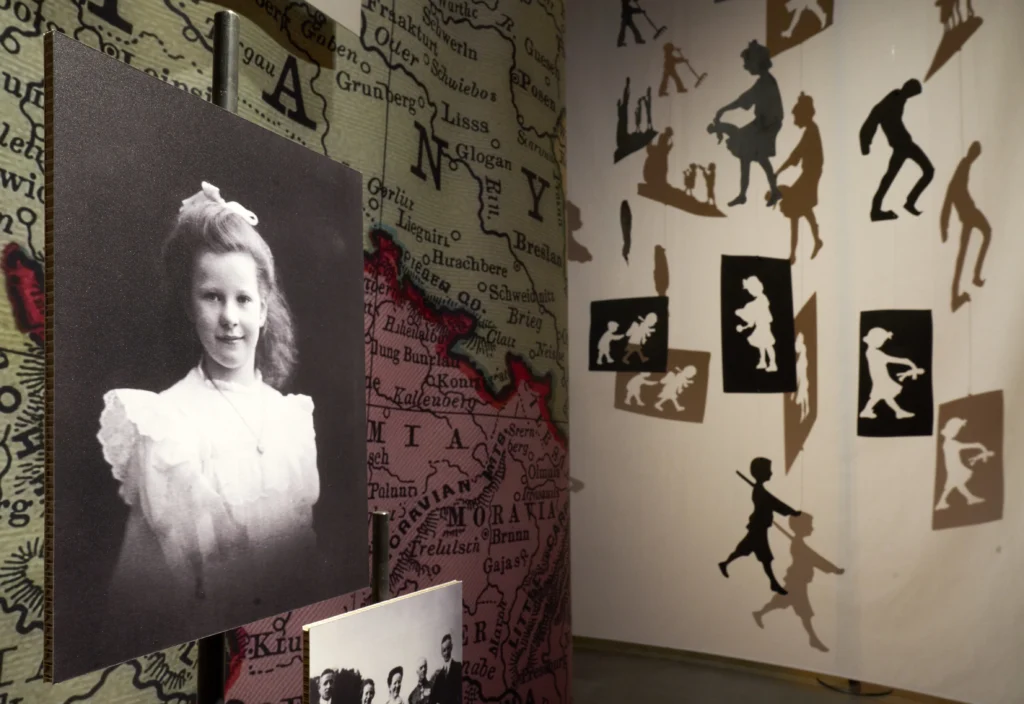 Detailansicht einer Ausstellung mit einem historischen Porträt eines Mädchens vor einer großformatigen Landkarte. Daneben hängen Scherenschnitt-Silhouetten spielender Kinder, die unterschiedliche Alltagsszenen darstellen.