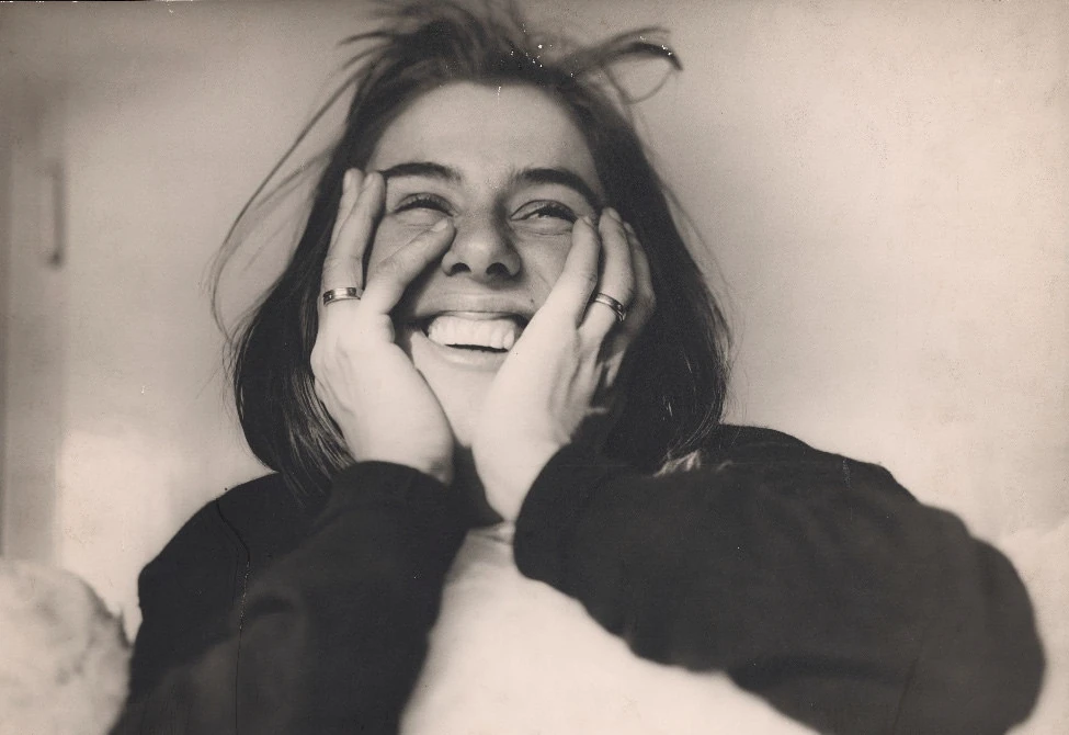 Auf einer schwarz-weiß Fotografie ist eine lachende junge Frau mit dunkelen Haaren zu sehen. Sie Trägt einen dunkelen Pullover und hält ihre Hände im Gesicht. Ihre Augen sind fast geschlossen, ihre Zähne sind beim Lachen zu sehen.