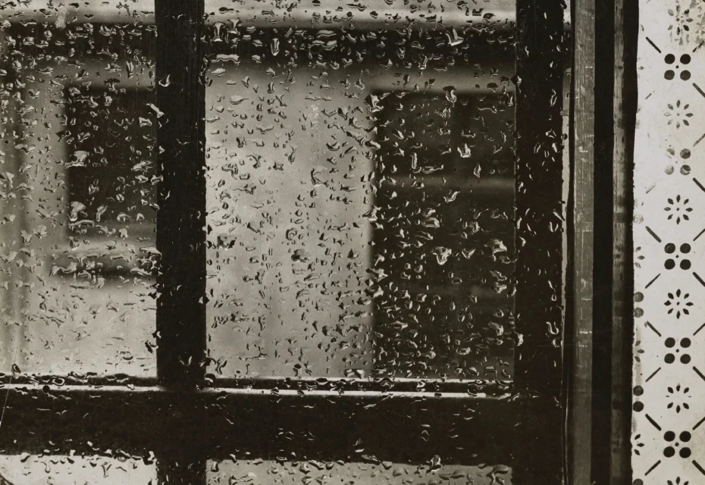 Eine schwarz-weiß Fotografie eines Fensters auf dem Regentropfen sind. Durch das Fenster sieht man verschwommen eine helle Hausfassade.