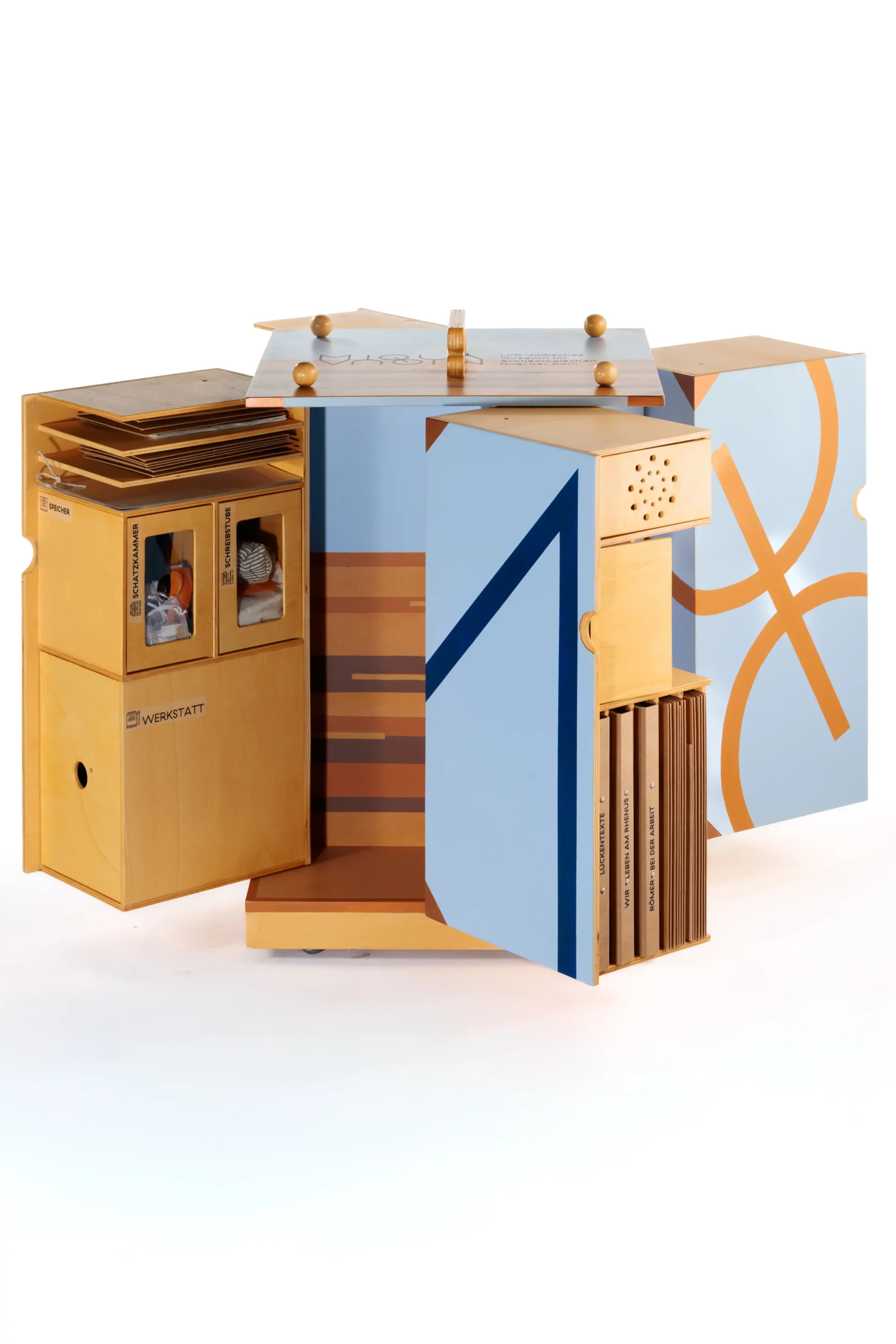Ein hölzernes, modular aufgebautes Möbelstück mit geöffneten Fächern und Schubladen. Es enthält verschiedene Materialien und Objekte und ist mit hellblauen sowie orangefarbenen grafischen Elementen gestaltet.