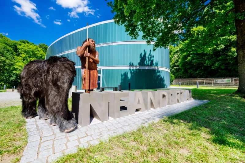 Zentral auf dem Bild sieht man den steinernen Schriftzug "Neanderthal". Links davon sieht man ein nachgestelltes Mammut mit schwarzem Fell und eine Neanderthaler Figur mit klassischer Kleidung und Stab. Im Hintergrund ist das Gebäude, oval mit blauen vertikalen Glasflächen, das Neanderthal Museum zu sehen.