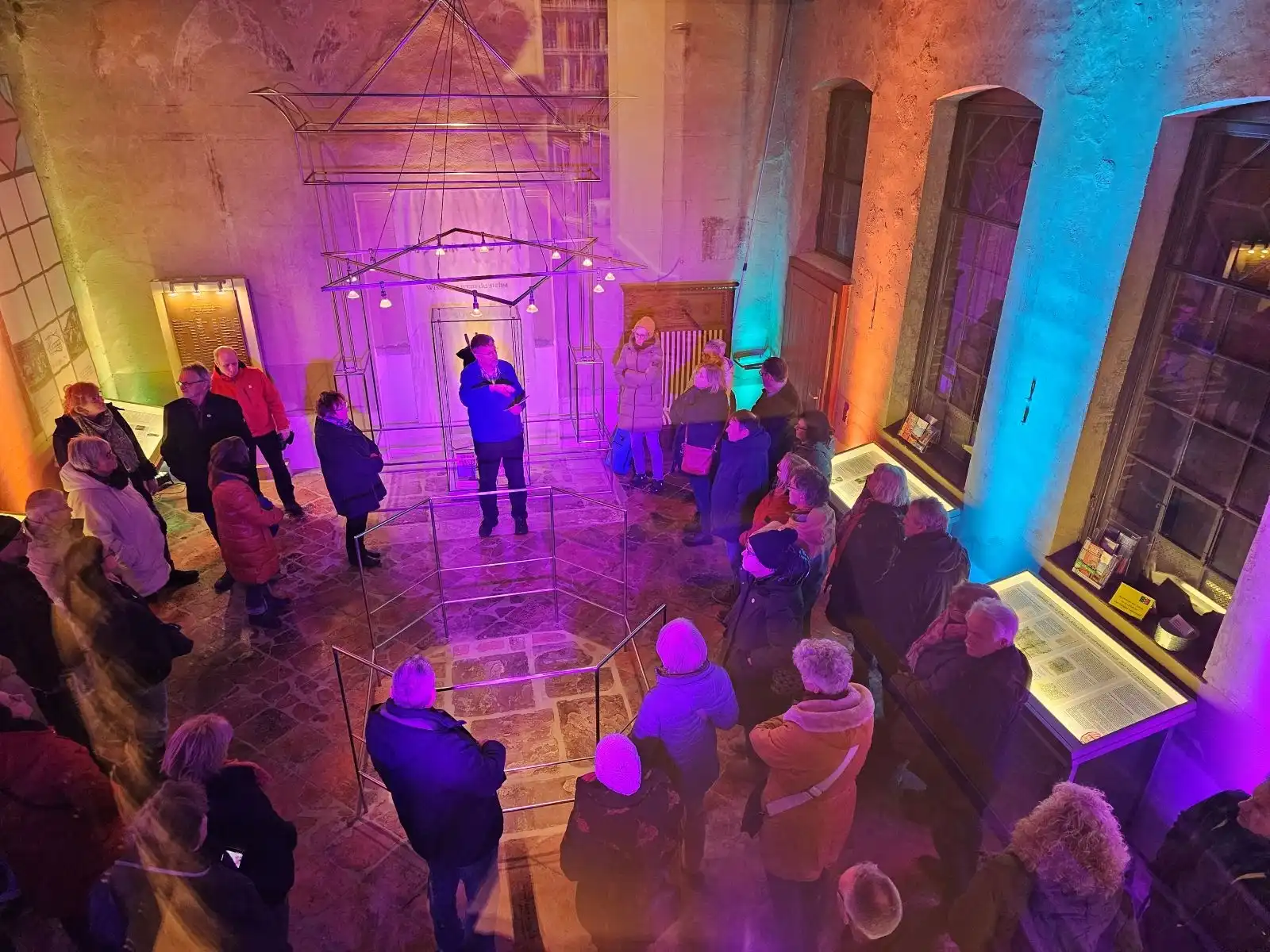 Mehrere Personen stehen in einem bunt Beleuchteten Ausstellungsraum in einem historischen Gebäude mit gemauertem Fußboden und hohen Fenstern. Der Blick ist von Oben auf die Gruppe. Zentral erklärt der Ausstellungsguide den Besuchern die Ausstellung.