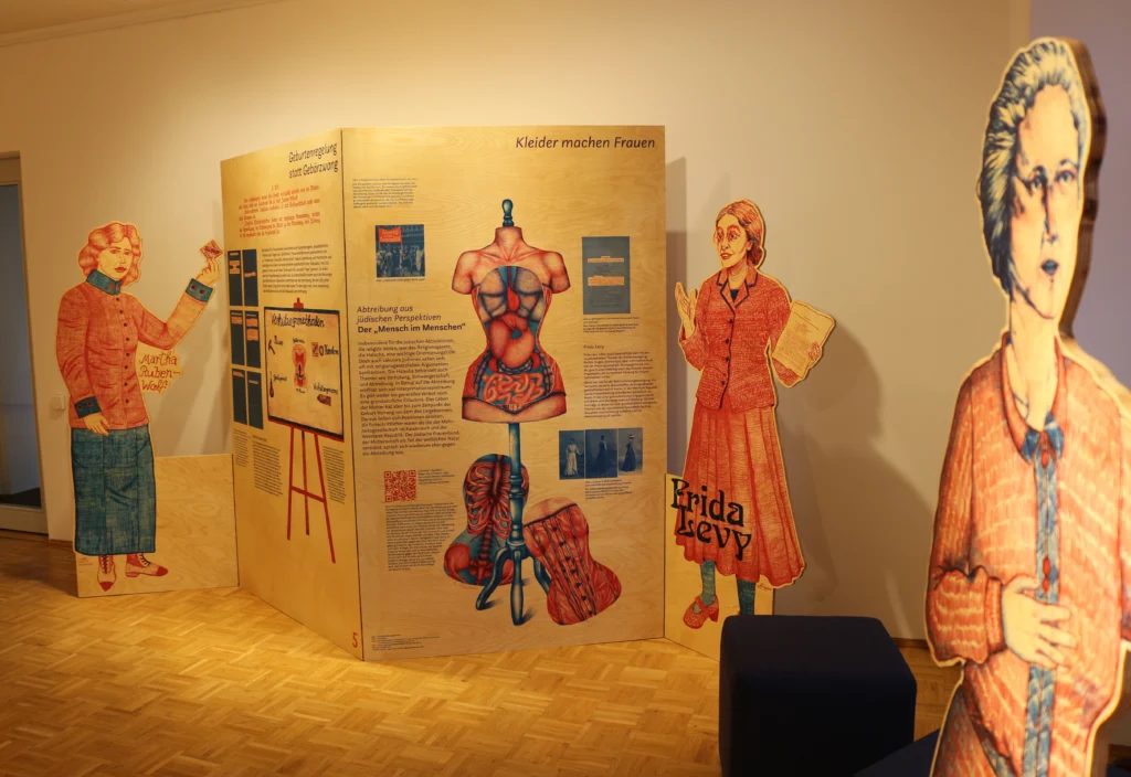 Blick in eine Museumsausstellung mit Informationstafeln im Hintergrund. Im Bild sind drei lebensgroße Figuren von Frauen als Aufsteller, und Informationstafeln zum Thema Geburtenregelung und Abtreibung im Bild. Die warm beleuchtete Ausstellung vermittelt einen historischen Kontext zur Frauenbewegung und jüdischen Aktivistinnen.