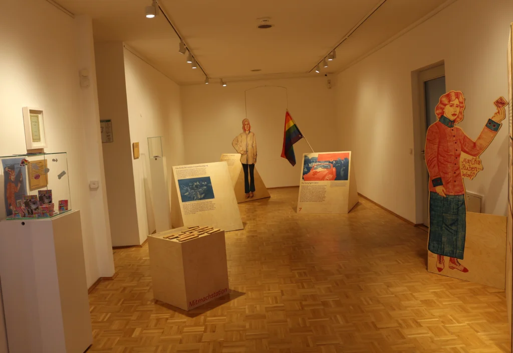 Blick in eine Museumsausstellung mit Informationstafeln im Hintergrund. Im Bild sind zwei lebensgroße Figuren von Frauen als Aufsteller, und Informationstafeln im Bild. Die warm beleuchtete Ausstellung vermittelt einen historischen Kontext zur Frauenbewegung und jüdischen Aktivistinnen.