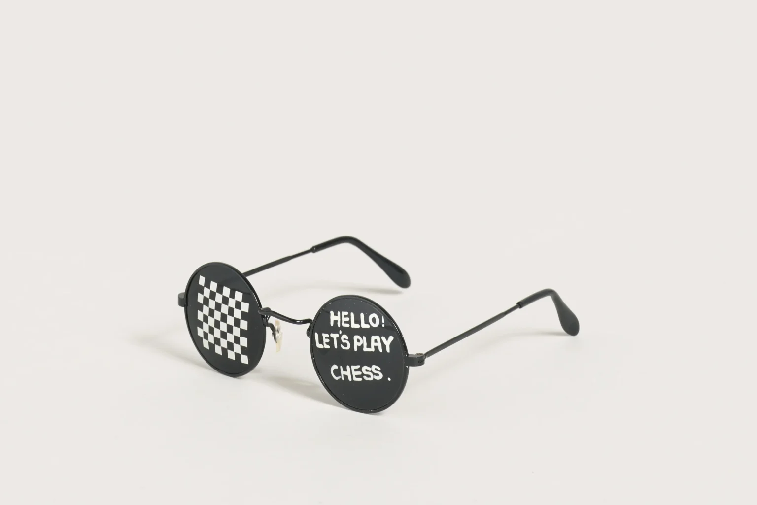 Auf einem hellbeigen Hintergrung ist mittig eine schwarze runde Sonnenbrille abgelegt. Auf dem linken Brillenglas ist ein Schachbrettmuster aufgemalt, auf dem rechten Brillenglas steht "Hello! Let's play chess."