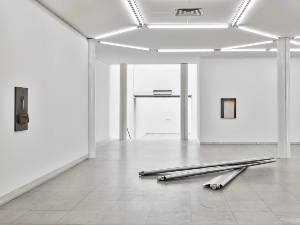 Heller, weitläufiger Ausstellungsraum mit weißen Wänden, Säulen und Deckenleuchten. An den Wänden hängen wenige abstrakte Kunstwerke. Auf dem grauen Fliesenboden liegen mehrere lange, zusammengerollte Metallbahnen.