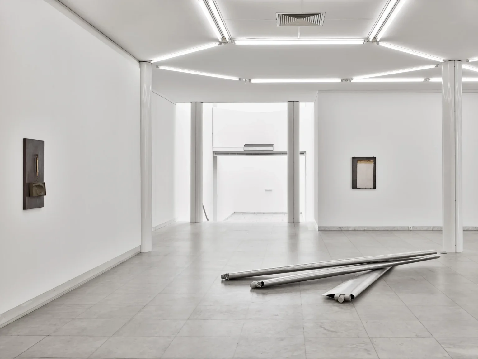 Heller, weitläufiger Ausstellungsraum mit weißen Wänden, Säulen und Deckenleuchten. An den Wänden hängen wenige abstrakte Kunstwerke. Auf dem grauen Fliesenboden liegen mehrere lange, zusammengerollte Metallbahnen.