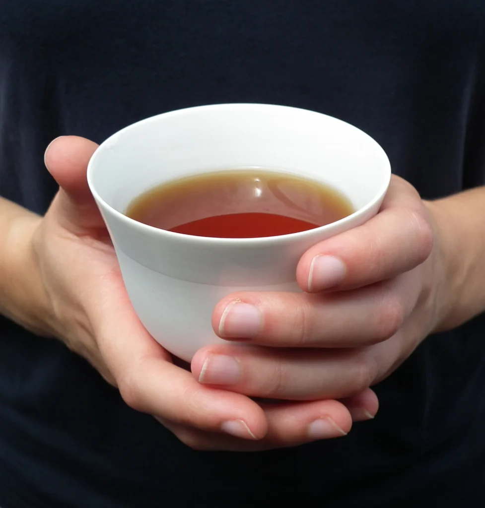 Zu sehen ist eine weiße, schlichte Keramik Teetasse, in der sich eine bernsteinfarbige Flüssigkeit befindet. Die Tasse wird von zwei Händen umschlossen. Der Hintergrund ist schwarz