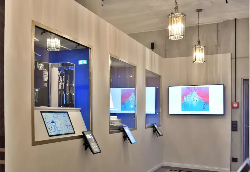 Innenansicht eines Ausstellungsraums mit beleuchteten Vitrinen und interaktiven Bildschirmen. In den Vitrinen sind historische Objekte ausgestellt; an der Wand zeigen mehrere Monitore farbige Darstellungen. Vor den Vitrinen befinden sich kleine Touchscreens.