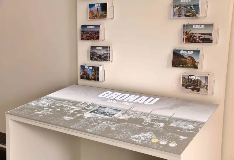 Zu sehen ist eine Ausstellnungsfläche, ein Tisch mit der Aufschrift "GRONAU 1946-1961" und einer schwarz-weiß Fotografie. Darüber sind in Plexiglashüllen Postkarten von unterschiedlichen Orten. Berlin, Hamburg, Gronau, New York, Hamburg, Duisburg und Düsseldorf