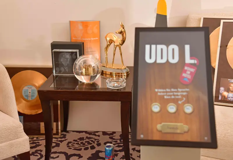 Im Vordergrund ein Aufsteller "UDO L."- im Hintergrund auf einem Tisch sind mehrere Musikpreise, u.A. Bambi zu sehen, an die Wand gelehnt mehrere goldene Schallplatten.