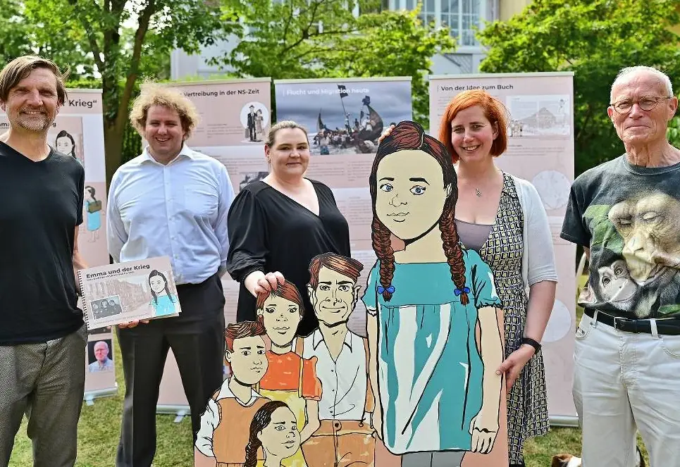 Fünf Erwachsene stehen bei einer Ausstellung im Freien vor Informationstafeln zur Geschichte einer Familie in der NS-Zeit. In der Mitte steht eine große gezeichnete Figur eines Mädchens mit Zöpfen; davor sind weitere gezeichnete Familienmitglieder zu sehen. Zwei Personen halten Informationsmaterial in den Händen.